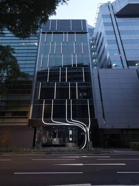 10 استراتژی برتر معماری پایدار برای ساختمانهای عمومی: راهنمای جامع 6 یک ساختمان مدرن با نمای شیشهای و نورپردازی منحصر به فرد شامل خطوط نورانی عمودی و طرحهای خمیده در ورودی.