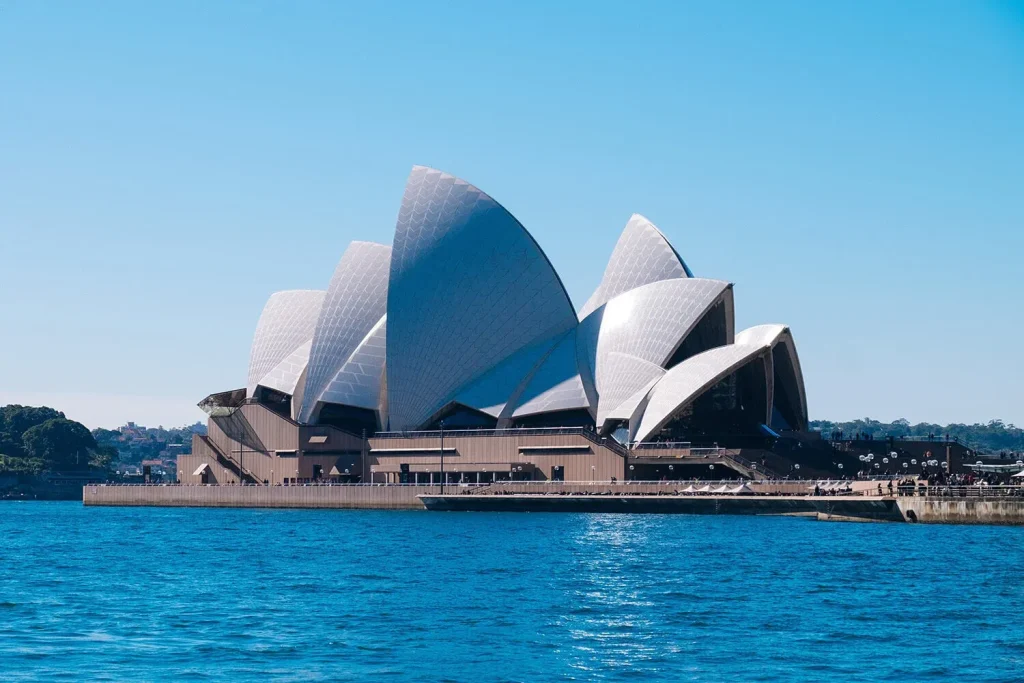 **خانه اپرای سیدنی، استرالیا، با سقف&zwnj;های سفید و صدفی شکل، در کنار آب.**(Sydney Opera House, Australia, with white, shell-shaped roofs, next to the water.)